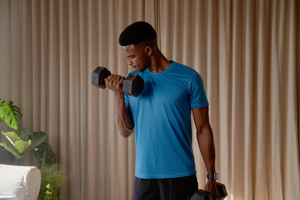 Front view of a Black African American young male exercising at home in his lounge lifting weights and dumbbells, bicep curls, keeping fit and healthy.High quality photo - Fotoğraf, Görsel