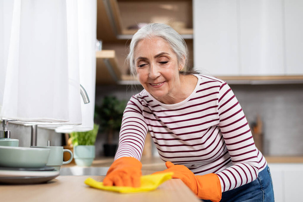 Портрет красивой старшей домохозяйки стиральный стол на кухне с тряпкой, улыбающаяся пожилая женщина в резиновых перчатках делает домашнюю работу, счастливая дама наслаждаясь уборка дома, крупным планом выстрел - Фото, изображение