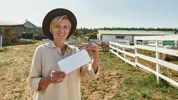 Adult caucasian female farmer holding and showing empty card on farm. White card for copy space. Smiling woman looking at camera. Modern countryside lifestyle. Agriculture and farming. Warm sunny day - Foto, immagini