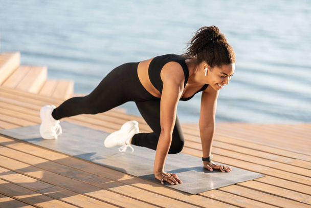 Athletic beautiful young black woman in sportswear training outdoors next to seaside, standing on yoga mat and stretching her body, listening to music, using wireless earbuds, copy space - Foto, Imagem