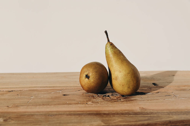Autumn, summer fruit still life. Two yellow pears on old rustic wooden table backgound in sunset light. Blurred beige wall, soft long shadows. Selective focus, farm food composition, copy space. - 写真・画像