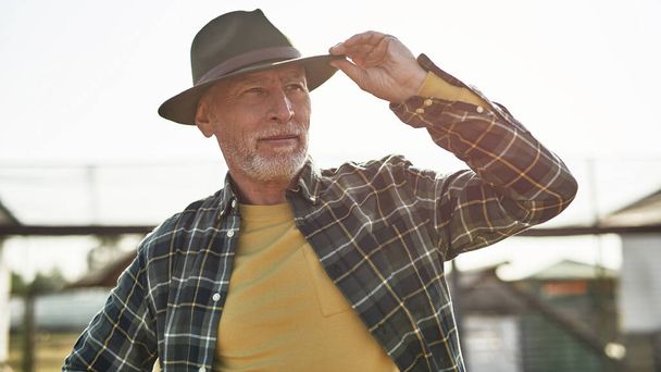 Mature male farmer looking away on blurred farm or ranch. Concentrated grey hair caucasian man wearing hat. Concept of modern countryside lifestyle. Agriculture. Warm sunny day - Foto, afbeelding