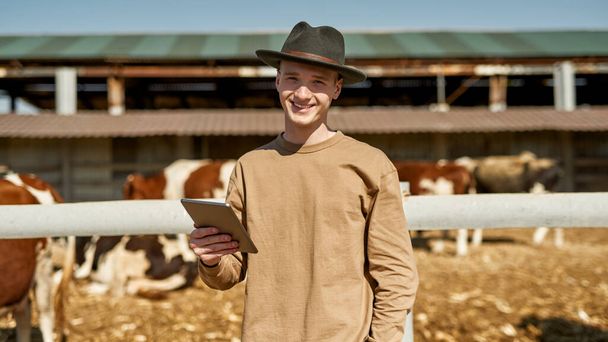 Front view of smiling male farmer with digital tablet near blurred paddock with milk cows on farm or ranch. Young caucasian guy looking at camera. Modern countryside lifestyle. Agriculture. Sunny day - Foto, immagini