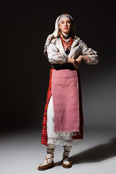 full length of ukrainian woman in dress with ornament and red beads standing on black  - Foto, immagini