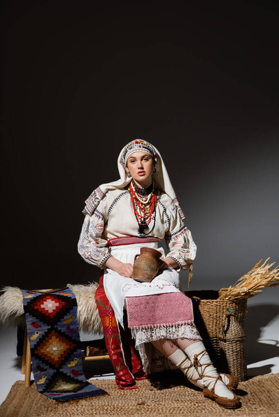 full length of young ukrainian woman in traditional clothing with ornament holding clay pot while sitting on bench on black - Foto, imagen