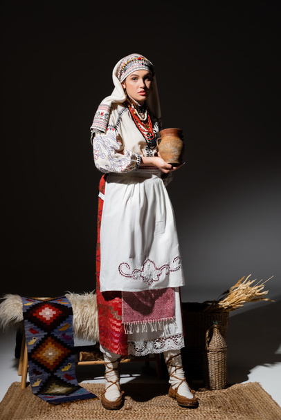 full length of pretty  ukrainian woman in traditional clothing with ornament holding clay pot while standing on black - Foto, Imagem