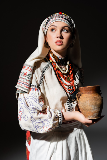 portrét krásné ukrajinské ženy v tradičním oblečení s ornamentem držení hliněný hrnec na černé - Fotografie, Obrázek