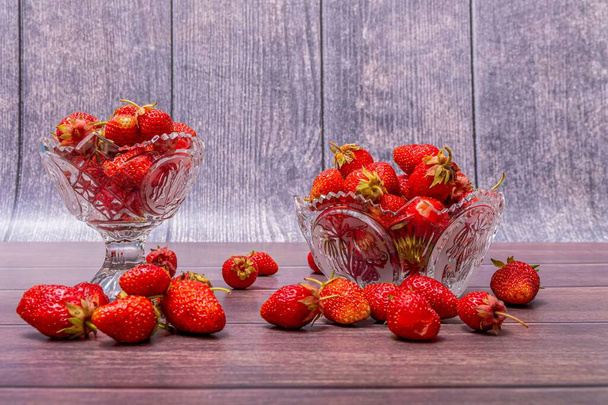 On the surface of a wooden table, there are two crystal vases with ripe red strawberries in them. - Φωτογραφία, εικόνα