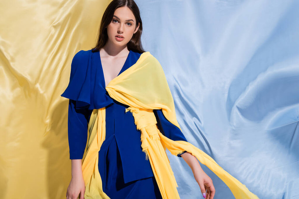 young ukrainian woman in color block clothing posing near blue and yellow fabric - Фото, изображение
