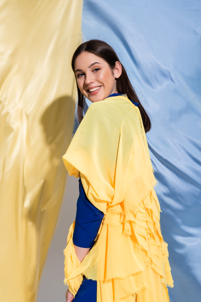 happy ukrainian woman in color block clothing posing near blue and yellow fabric - Photo, Image