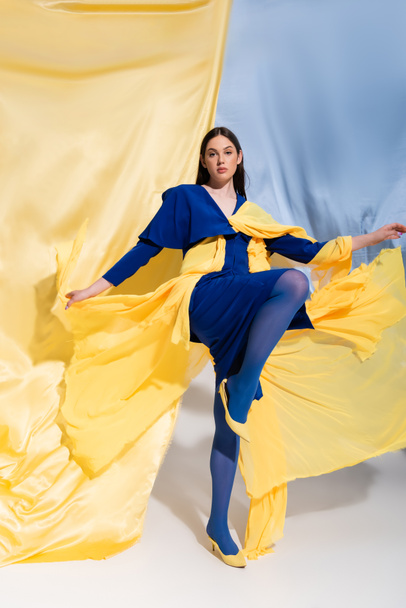 full length of young ukrainian woman in stunning color block clothing posing near blue and yellow fabric - Fotografie, Obrázek