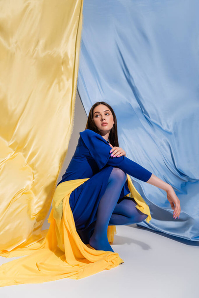 trendy young ukrainian woman in stylish color block clothing sitting near blue and yellow flag - Valokuva, kuva