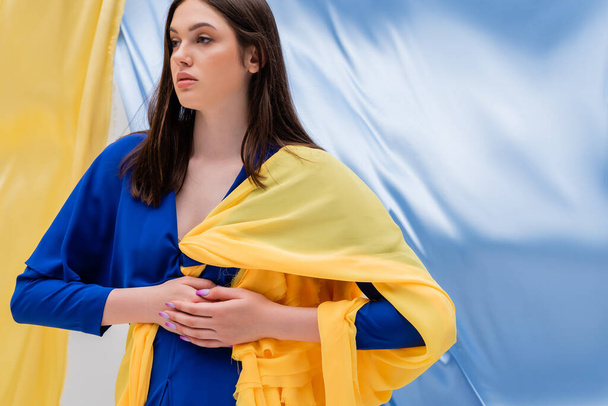 patriotic ukrainian young woman in stylish clothing posing near blue and yellow flag  - 写真・画像