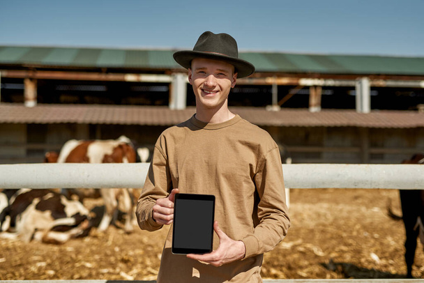 Smiling male farmer showing digital tablet with blurred milk cows in paddock on background on farm. Young caucasian guy looking at camera. Modern countryside lifestyle. Agriculture. Sunny day - Fotoğraf, Görsel