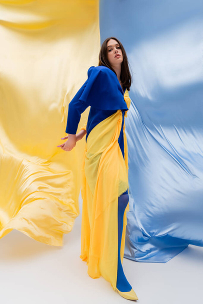 full length of patriotic ukrainian young woman in stylish clothing posing near blue and yellow fabric - Фото, изображение