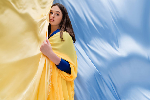 mujer joven ucraniana patriótica con ojos azules posando cerca de tela azul y amarilla - Foto, imagen