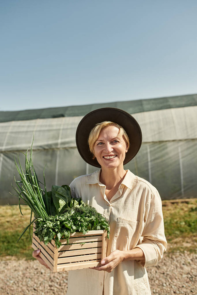 Smiling adult blonde caucasian female farmer holding wooden basket with organic greens near greenhouse in village or farm. Fresh eco harvest. Modern cauntryside lifestyle. Agriculture. Sunny day - Фото, зображення