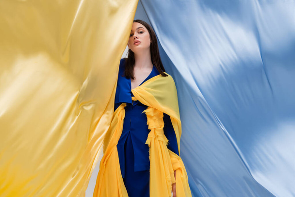 patriotic ukrainian young woman in fashionable clothing posing near blue and yellow fabric  - Φωτογραφία, εικόνα
