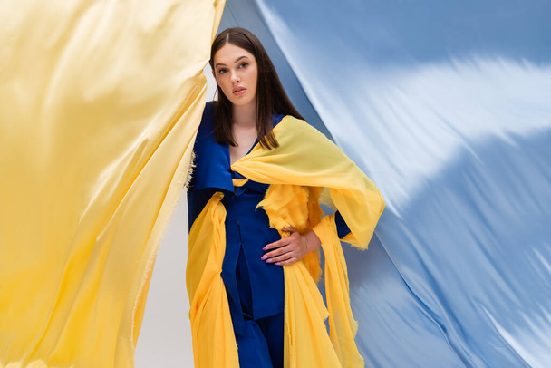 patriotic ukrainian young woman in trendy clothing posing with hand on hip near blue and yellow fabric  - Foto, imagen