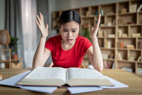 Big mistake. Amazed unhappy teenager chinese girl reading book, gesticulate in room or library interior. Human emotions, shocked facial expression, prepare for exam at home during covid-19 outbreak - Foto, imagen