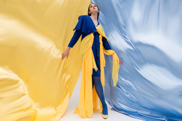 plná délka vlasteneckého ukrajinského modelu v módním oblečení pózuje v blízkosti modré a žluté tkaniny - Fotografie, Obrázek