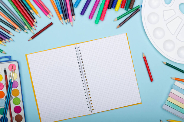 Blank sheet of paper of an open math notebook, art school supplies for painting, on a blue background with copy space for text. Colorful pencils, markers, paints, crayons. Top view. Back to school - Photo, image