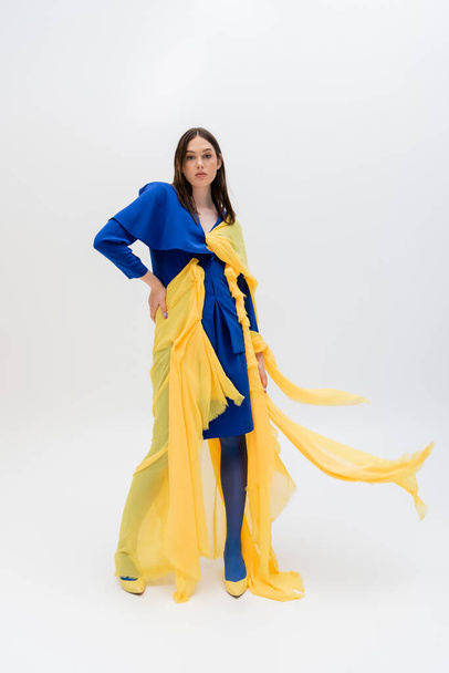full length of stylish ukrainian woman in blue and yellow outfit posing with hand on hip on grey - Fotografie, Obrázek