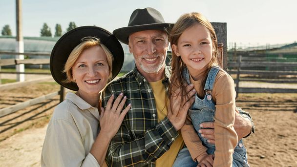 Portrait of smiling caucasian grandparents and granddaughter looking at camera on blurred farm. Farmer family spending time together. Modern countryside lifestyle. Agriculture and farming. Sunny day - Foto, Bild