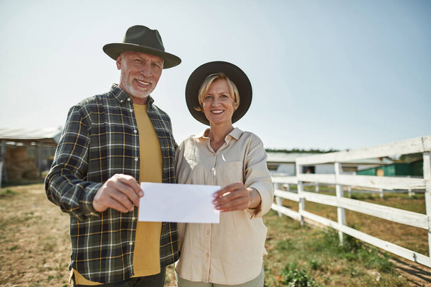 Mature caucasian farmer couple hugging and showing empty card on farm. White card for copy space. Happy woman and man looking at camera. Modern countryside lifestyle. Agriculture. Warm sunny day - Foto, immagini