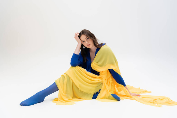 patriotic ukrainian young woman in blue and yellow clothes sitting while posing on grey - Foto, imagen