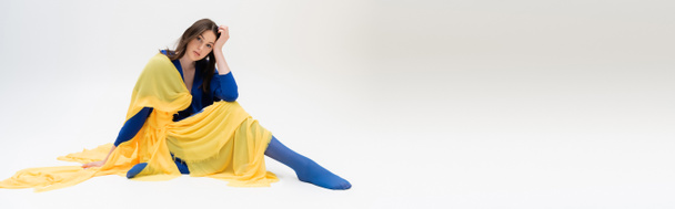 patriotic ukrainian young woman in blue and yellow clothes sitting while posing on grey, banner - Photo, image