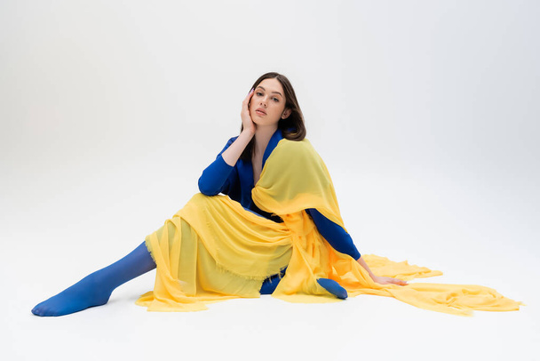patriotic ukrainian young woman in blue and yellow outfit with tights sitting while posing on grey - Фото, зображення