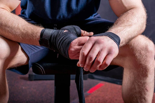 Cropped body of strong young mixed martial artist who sits on couch and wraps elastic bandage to fix his wrist for open fist fight. - Foto, afbeelding