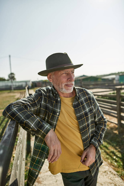 Mature male farmer looking away at wooden fence on farm or ranch. Concentrated caucasian man wearing hat. Concept of modern countryside lifestyle. Agriculture and farming. Warm sunny day - Fotó, kép