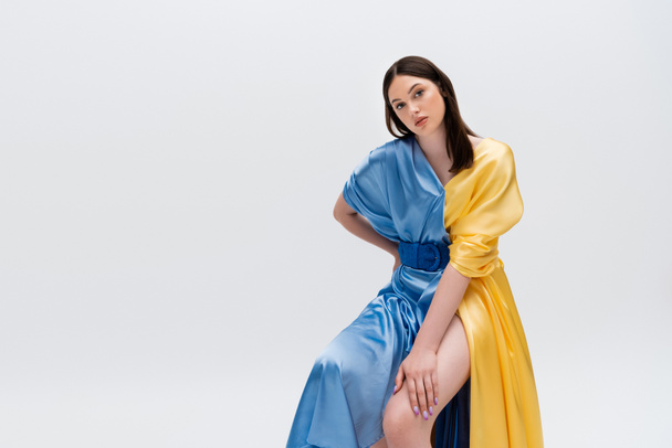 sensual young ukrainian woman in blue and yellow dress posing with hand on hip while sitting isolated on grey - Fotoğraf, Görsel