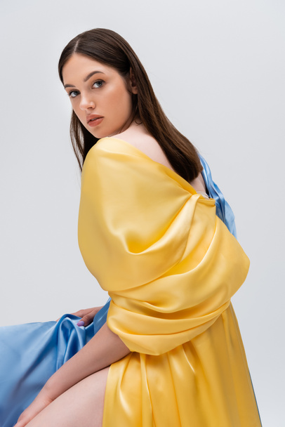 young ukrainian woman in blue and yellow dress posing while sitting isolated on grey - Foto, immagini