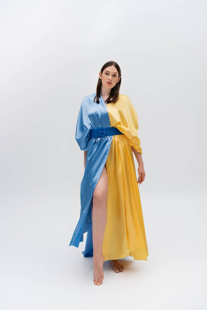 full length of barefoot young ukrainian woman in blue and yellow dress posing on grey - Fotó, kép