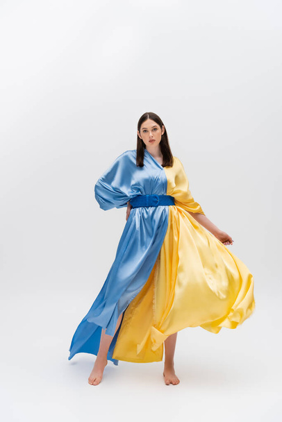 full length of barefoot young ukrainian woman in blue and yellow dress posing with hand on hip on grey - Фото, изображение