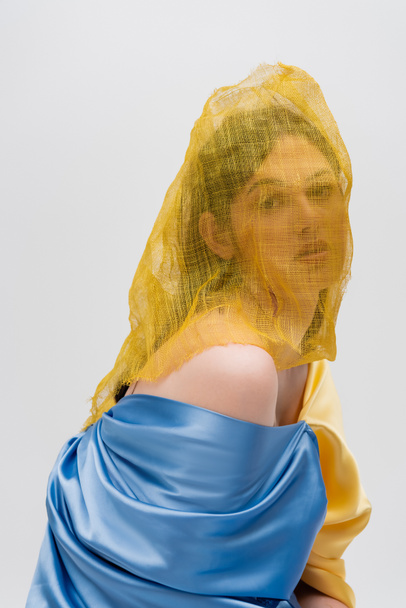 young ukrainian woman woman with yellow drapery covering face looking at camera isolated on grey - Foto, immagini