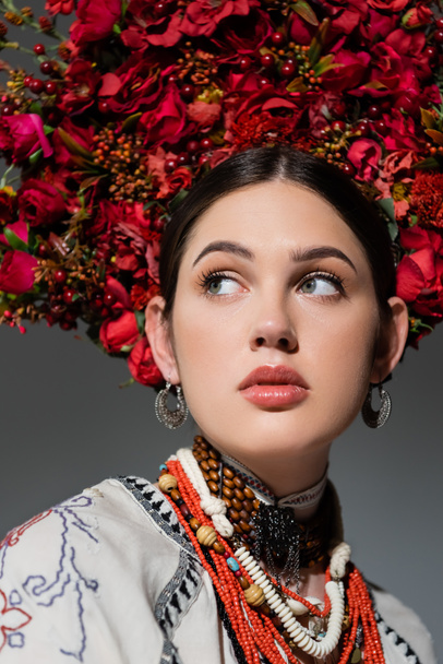 portrait of pretty ukrainian woman in traditional clothing and floral red wreath looking away isolated on grey - Photo, Image