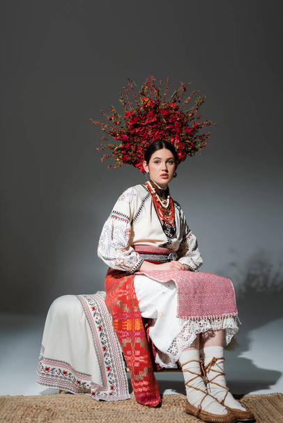повна довжина красивої української жінки в традиційному одязі і червоний вінок з квітами і ягодами на сірому
 - Фото, зображення
