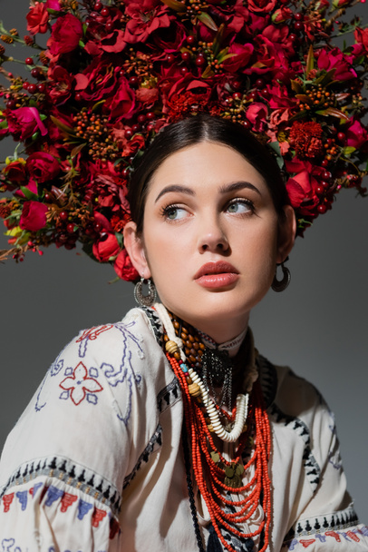 portrait of ukrainian woman in traditional clothing and floral red wreath looking away isolated on grey - Foto, Bild