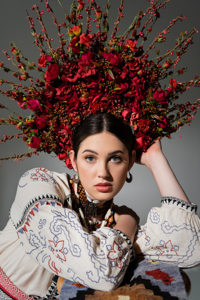 portrait of ukrainian woman in traditional clothes and floral wreath with red berries looking at camera isolated on grey - Foto, Imagen