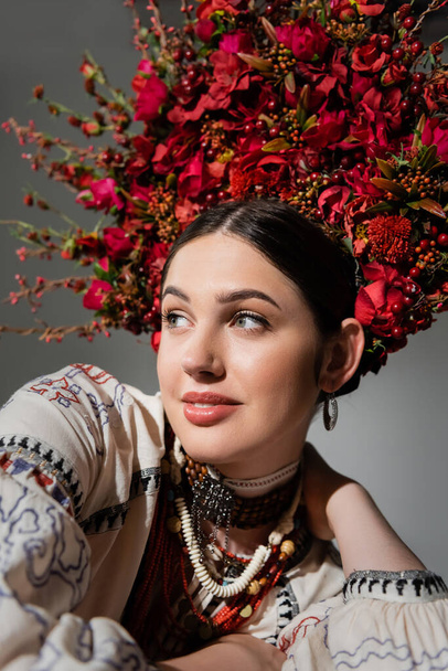 portrait of smiling ukrainian woman in traditional clothes and floral wreath with red berries isolated on grey - Foto, Imagem