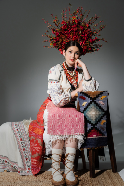 thoughtful ukrainian woman in traditional clothes and red wreath with flowers and berries on grey - Foto, afbeelding