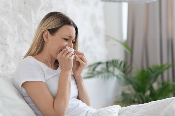 Ill unhappy blonde millennial lady in pajamas sitting in bed at home, sneezing, using napkin, suffering from pain. Cold, flu or coronavirus concept, panorama with copy space, closeup - Fotó, kép