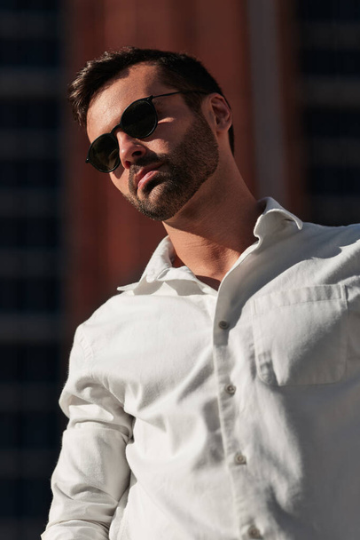 Self assured young bearded Middle Eastern male model with dark hair in elegant white shirt and sunglasses standing on city street on sunny day - Foto, afbeelding