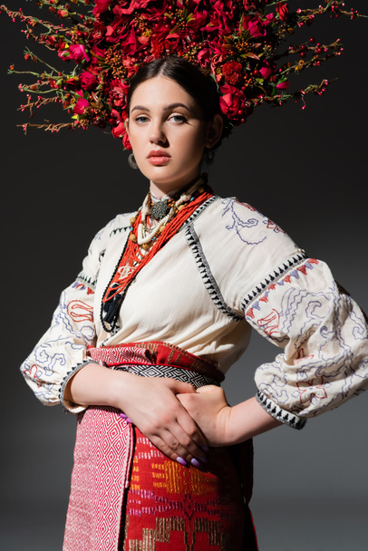 brunette and pretty ukrainan woman in floral wreath with red berries posing with hand on hip on dark grey - Photo, Image