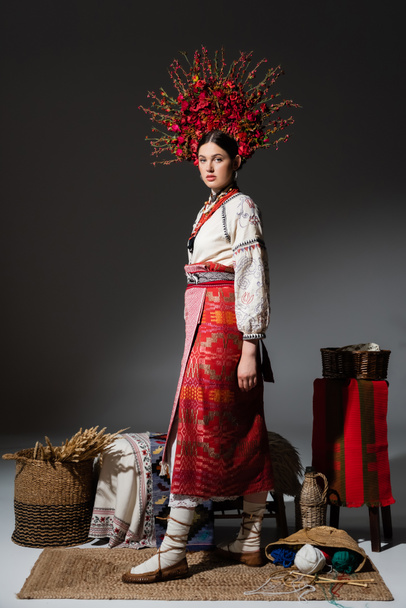 full length of ukrainian woman in traditional red floral wreath standing near yarn balls and wheat on dark grey - Foto, Imagem