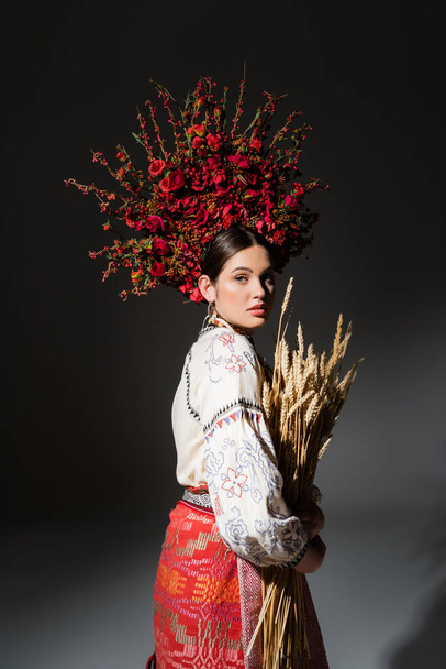brunette and young ukrainan woman in floral wreath with berries holding wheat on black - 写真・画像